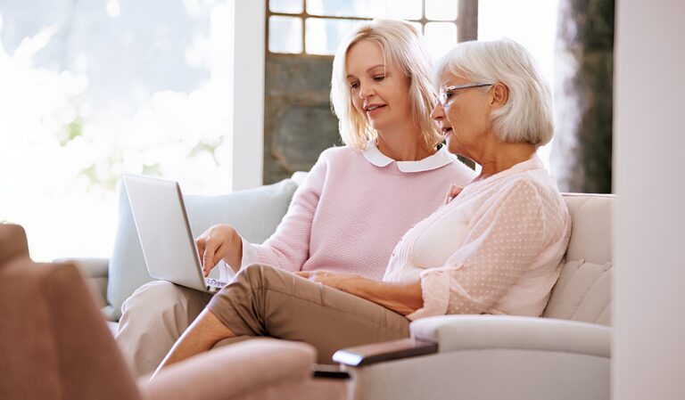 Two elderly women sit together on a light sofa, looking at an open laptop on one’s lap in a bright living room.