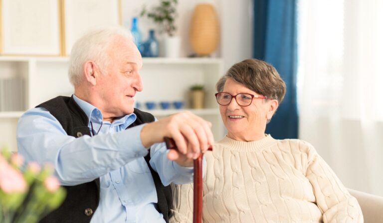 An elderly man and woman sit close together on a sofa, smiling at each other while the man rests his hand on a wooden cane in front of him.