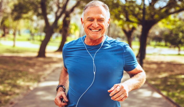 Smiling middle-aged man jogging on a sunlit park path in a blue athletic shirt with white earbuds in.
