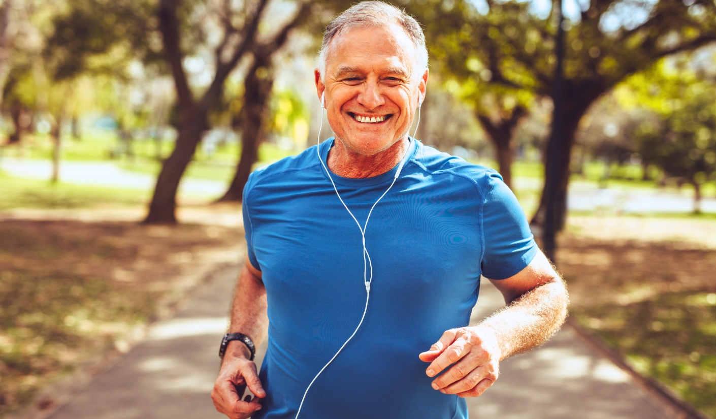 Smiling middle-aged man jogging on a sunlit park path in a blue athletic shirt with white earbuds in.