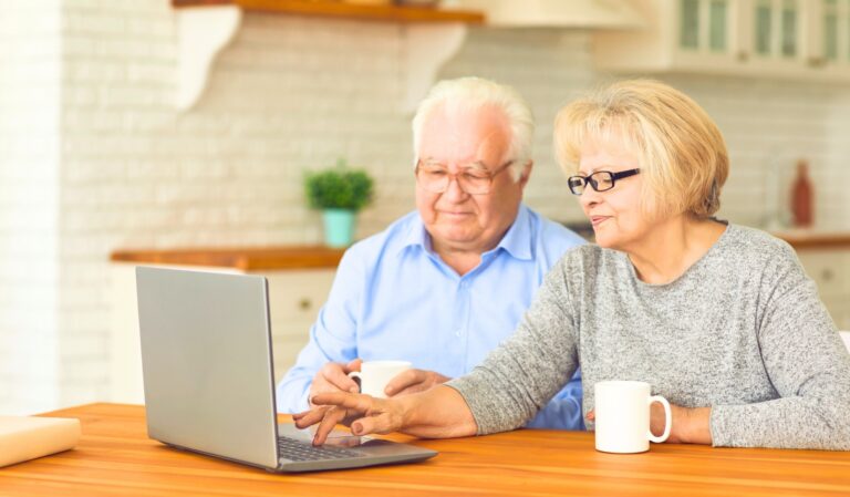 Elderly couple at a kitchen table, focused on a laptop with coffee mugs in hand.